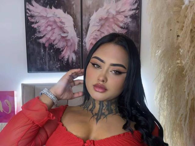 LairaHearvy with black hair, red dress, tattoo on neck, pink wing art, and silver bracelet, looking at camera with hand near face, full lips, and winged eyeliner.