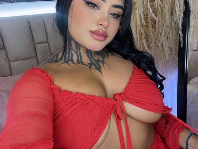 LairaHearvy with black tattoos on her neck, wearing a red, sheer top that reveals her big tits, has long black hair, and is looking at the camera.