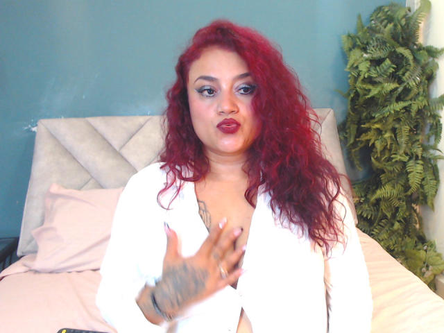 VictoriaBlake with red curly hair, white robe, tattoos on hand, pouting with lipstick, slightly open robe showing cleavage, right hand raised, green wall, fern plant in background.