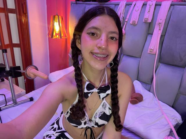 NamySex mit geflochtenem, braunem Haar, trägt einen schwarz-weißen Bikini mit Kuhmuster, lächelt und hat eine Nasenring. Sie liegt auf einem Bett mit einem Dildo und BDSM-Fesseln..