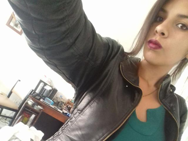 VioletaStone with pink lipstick, wearing a black leather jacket and green top, raising her arm for a selfie. Her tits are partially visible under the jacket.