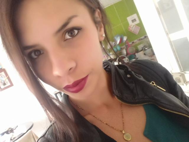 VioletaStone has light skin, brown eyes, and dark brown hair. She's wearing a black jacket, green top, and red lipstick with a gold necklace. Her tits are visible.