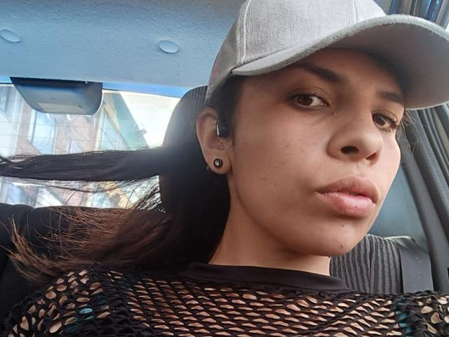 VioletaStone, wearing a white cap, black mesh top, and ear gauges, has puffy lips and brown eyes, with dark hair flowing from under the cap, in a car.