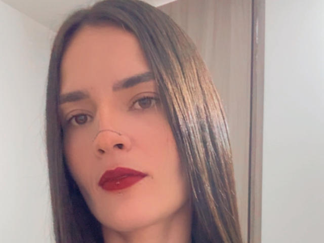 EmmaHerby with straight brown hair, dark red lipstick, nose ring, and thick eyebrows, looking into the camera with a slight smile.