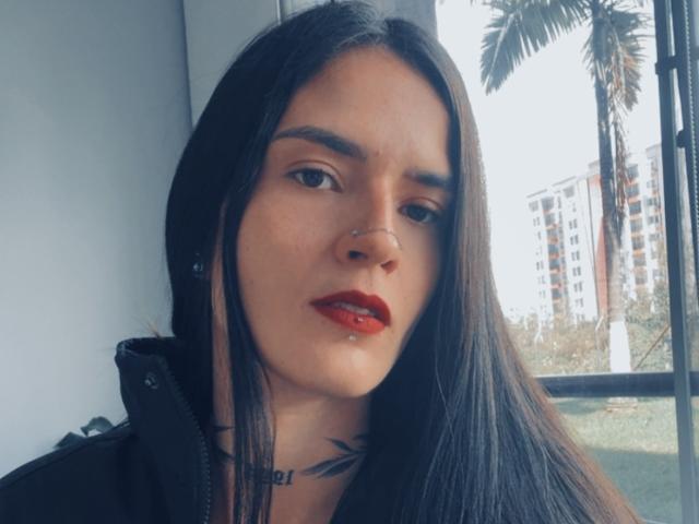 EmmaHerby has long black hair, red lipstick, and a nose piercing. She's looking slightly to the side with a subtle, confident expression. Her tits are barely visible.