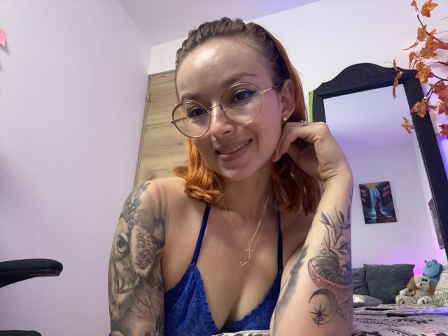SarahRousen with glasses, orange hair, tattoos on arms, blue lace bra, smiling, holding cheek, small tits, necklace, sitting, mirror on right, orange leaf decor, soft lighting.