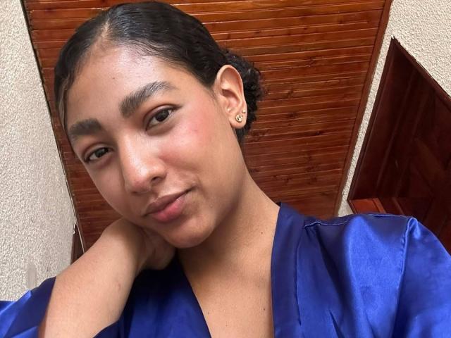 NatalyBlonder has dark hair in a bun, brown eyes, and wears a blue satin robe. Her face is slightly flushed, with a subtle smile and her hand on her neck.