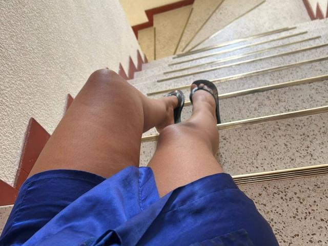 NatalyBlonder's legs in blue shorts, wearing black flip-flops, standing on a speckled stone staircase, looking upward. Her brown skin is visible, showing a slight knee crease.