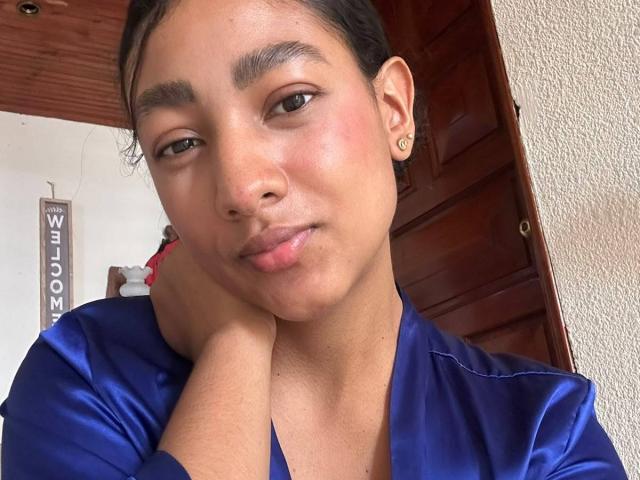 NatalyBlonder, con la pelle marrone, il naso piccolo, le sopracciglia scure, un trucco leggero e un sorriso carino. Indossa una camicia blu lucida e appoggia il mento sulla mano..