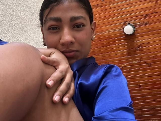 NatalyBlonder, con i capelli neri corti, la pelle marrone e gli orecchini dorati, tiene le sue grandi tette nude e marroni, vestita con una camicia blu, e guarda la fotocamera con un leggero sorriso..