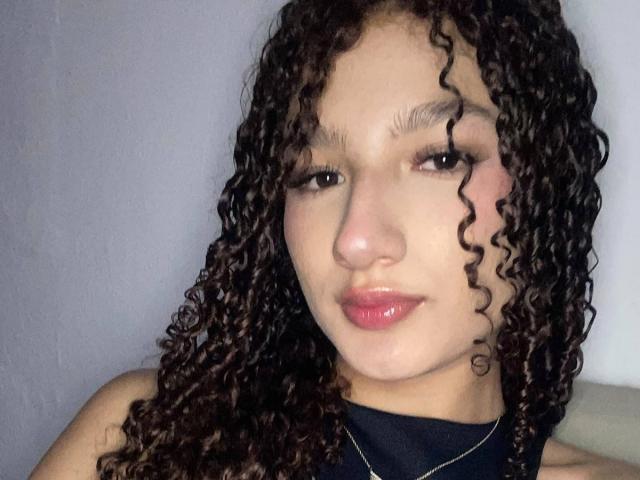 Nathacha Evans, con cabello castaño rizado, tono de piel medio, labios rosados brillantes, una blusa negra y un delineador de ojos sutil, mirando ligeramente hacia la derecha, y llevando un collar delgado..