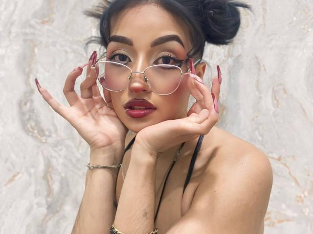 LunaixMoon, com cabelo escuro preso em coques, usando óculos, batom vermelho e unhas longas, segurando o rosto com as mãos, exibindo seus pequenos seios, em frente a uma parede branca..