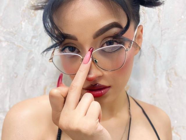 LunaixMoon, avec des lunettes, des ongles roses et du rouge à lèvres rouge, touchant son nez avec son doigt, cheveux foncés attachés en chignons, haut en bandeau noir, regardant la caméra, lumière douce, expression confiante..