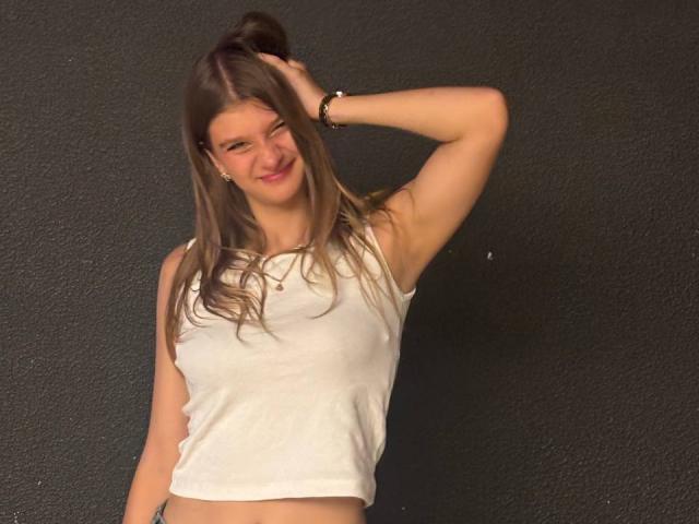 HilariaTabar with long brown hair, smiling, wearing a white sleeveless crop top, right arm raised, touching her head, showing her bare midriff, black textured wall in background.
