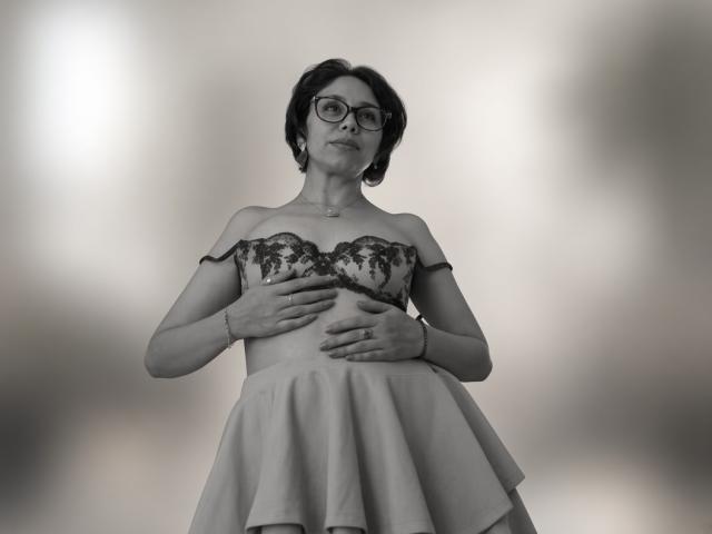 StephanieSun with glasses, dark short hair, off-shoulder lace dress, hands on her tits, standing, wearing a ring, necklace, and bracelet, black-and-white photo.