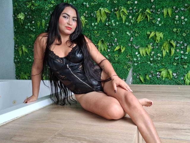 Dayana Alarcon mit langen, schwarzen Haaren, trägt einen schwarzen Overall aus Spitze und Leder, sitzt auf einem Holztisch, die Beine gekreuzt, die Hände auf dem Tisch, im Hintergrund eine grüne, strukturierte Wand..