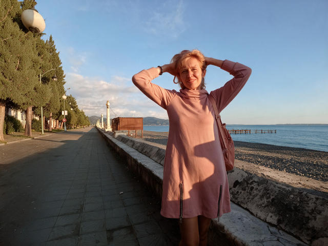 TrinaAdair, com as mãos no vestido rosa, em pé em uma trilha à beira da praia, sorrindo, segurando o cabelo, bolsa rosa no ombro, céu azul, água calma, píer de madeira, poste de luz, árvores à esquerda..
