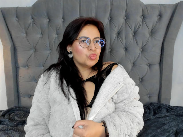 IvonnaLune, con i capelli scuri, gli occhiali blu e il rossetto rosa, indossa un morbido accappatoio grigio, un reggiseno di pizzo nero e orecchini di diamanti, con le labbra leggermente schiuse, seduta su un letto..