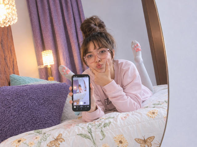 IsabelaP with glasses, pigtails, pink pajama top, white socks with pink paw prints, holding phone showing her face, lying on floral bed, pouty expression, mirror reflection.