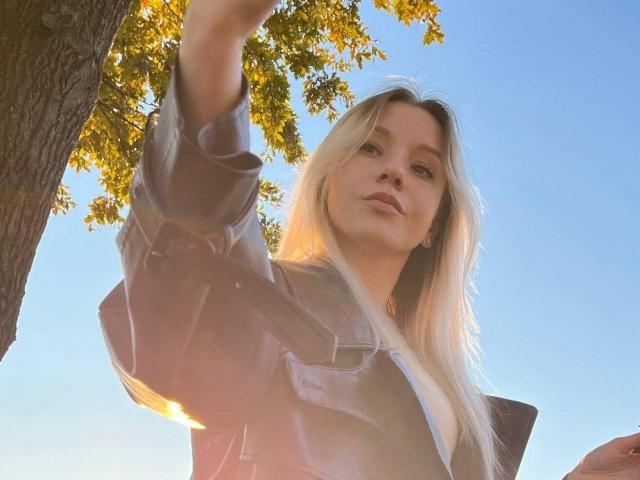 CherelleBettle with long blonde hair, wearing a translucent shirt, raising her arm, sunlight through leaves, clear blue sky, her face slightly tilted, looking downward.
