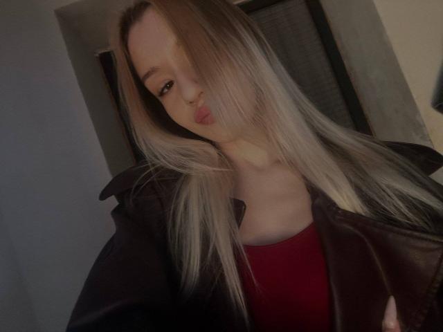 CherelleBettle with long blonde hair, pouty lips, wearing a black leather jacket over a red top, has a pale skin tone, and a subtle, sexy look.