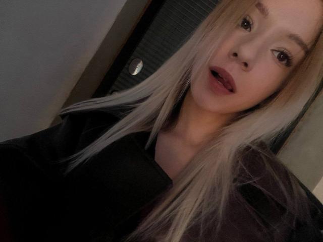 CherelleBettle with long blonde hair, brown eyes, and slightly open mouth, wearing a black shirt, in a dimly lit room, looking at the camera with a subtle smile.