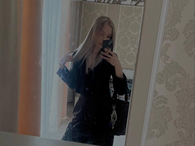 CherelleBettle with long blonde hair, wearing a black dress, taking a mirror selfie with a phone in a dimly lit room, holding her hair with her right hand.
