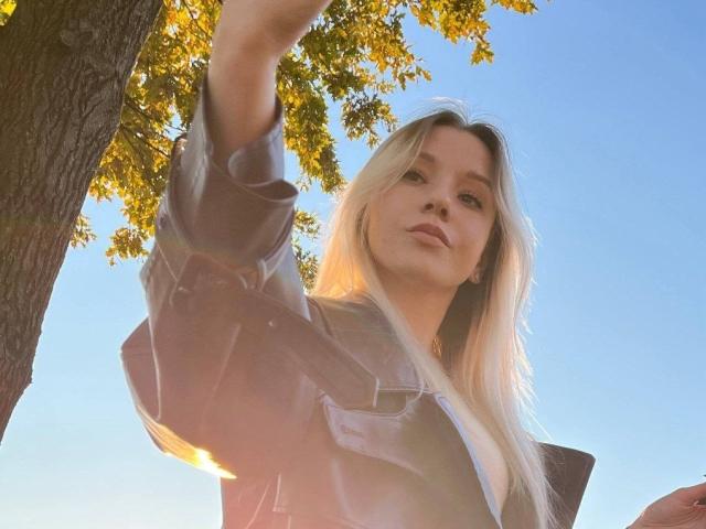 CherelleBettle with long blonde hair, wearing a shiny black jacket, taking a selfie from a low angle, sunlight filtering through tree leaves, clear blue sky in background.