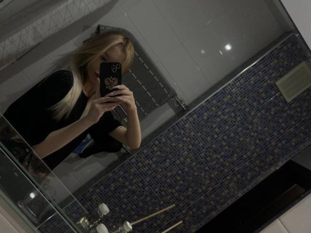 CherelleBettle with blonde hair, black top, taking a mirror selfie in a bathroom with blue mosaic tiles, holding a black phone, visible breasts, and a toilet in the background.