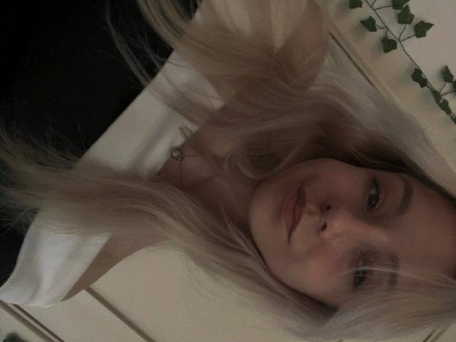 CherelleBettle with long, blonde hair lying down, showing her face and small nose. She has light skin and a subtle smile, wearing a white shirt.