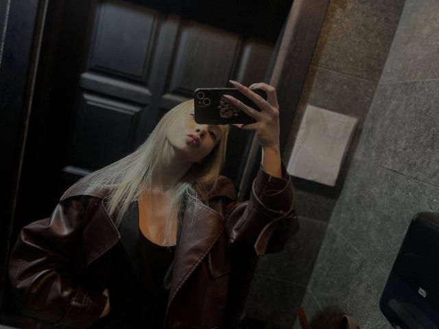 CherelleBettle with blonde hair, wearing a brown leather jacket over a black top, taking a mirror selfie with a black phone, pouty lips, and a confident pose.