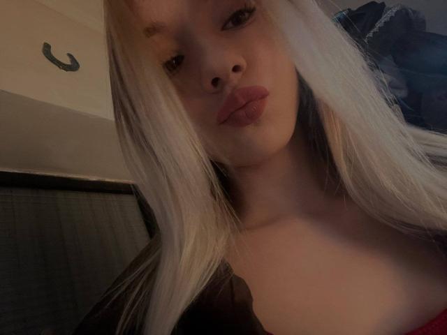 CherelleBettle with blonde hair, pouty lips, and fair skin, showing her tits in a low-light selfie. Her brown eyes are slightly open, and she's wearing a red top.