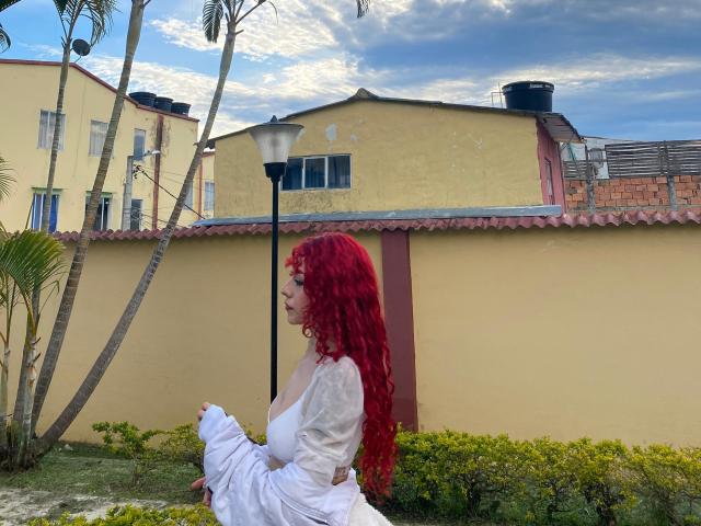 GaiaDoll with red curly hair, white dress, and visible tits, sitting outside, holding her dress, under a lamppost, with a yellow building and palm trees in the background.