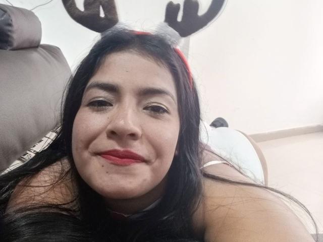 ArianithaXox with long black hair, red lips, and reindeer antlers, lying on a brown couch, smiling at the camera, showing her chubby face and small nose.