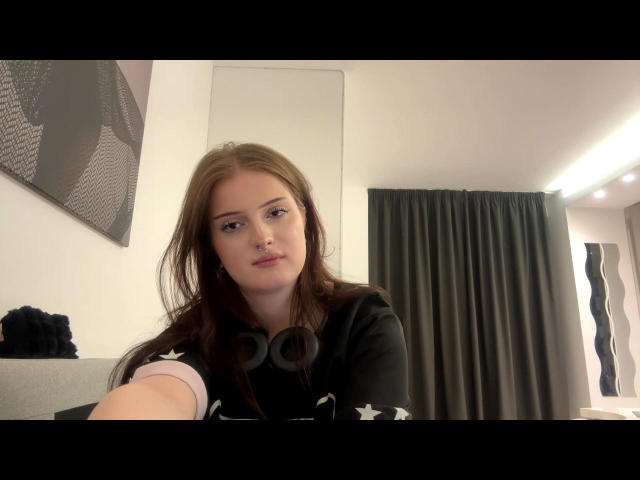 Kaleigh Diprima, met bruin haar, een zwart shirt en een koptelefoon, kijkt naar de camera, met subtiele make-up, een zachte uitdrukking, lichte huid, gemiddeld postuur, binnen, een selfie, een lichte glimlach, ontspannen..