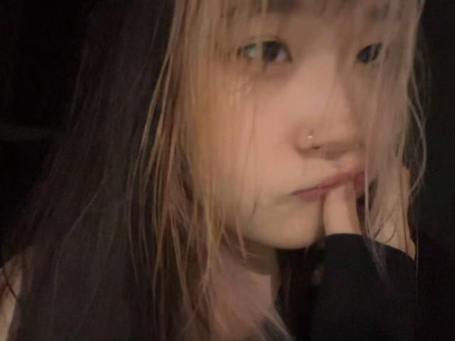 JungMcgranor with blonde and dark brown hair, nose piercing, finger near mouth, black top, slightly blurred, looking up with subtle smile.