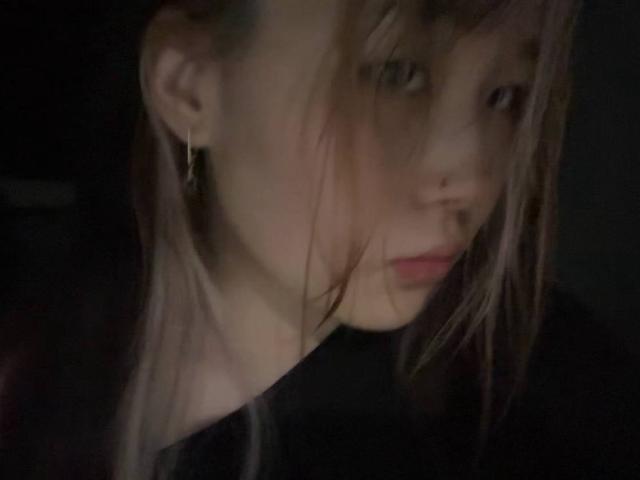 JungMcgranor has pale skin, a nose ring, and small earrings, with messy brown hair covering one eye, wearing a black shirt, in a dimly lit photo.