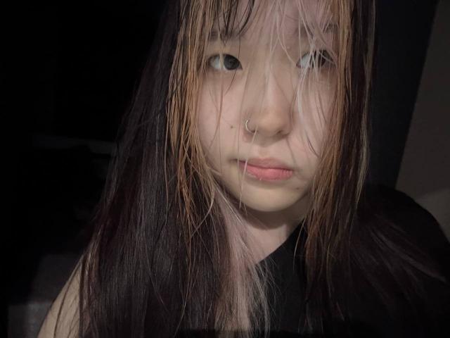 JungMcgranor with messy black and blonde hair, nose ring, pouty pink lips, and slightly tired eyes, wearing a black shirt, in dim lighting.