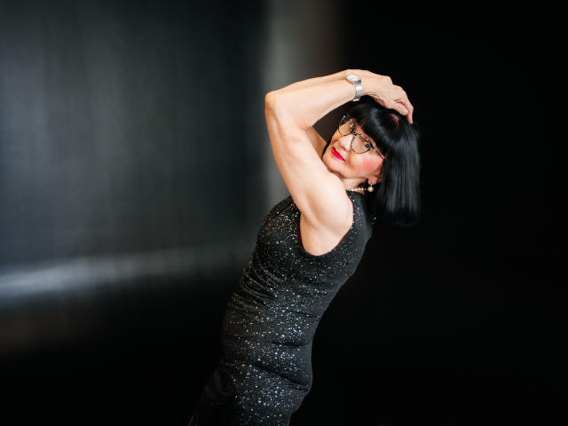 TiniaMartin with black bob, wearing sparkly black dress, arms raised, showing muscular arms, red lipstick, black background, confident, sexy, looking over shoulder, wearing silver bracelet.