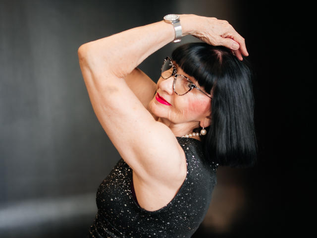 TiniaMartin with black bob, glasses, red lipstick, sparkly sleeveless top, pearl necklace, silver watch, arms raised, smiling, showing toned arms and chest, black hair, sexy.