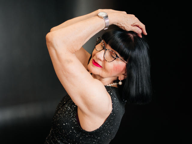 TiniaMartin with black bob, glasses, red lipstick, black sparkly sleeveless dress, arms raised, showing toned arms and small, saggy tits, wearing a silver watch and earrings.