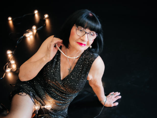 TiniaMartin, a middle-aged woman with black bob and glasses, wearing a sparkly black dress, holding a pearl necklace, sitting with string lights, showing her tits and ass.