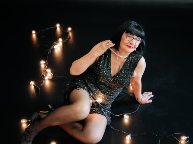 TiniaMartin with black bob, glasses, red lipstick, wearing a sparkly black dress, pearl necklace, and black high heels, sitting on black floor with string of lights.