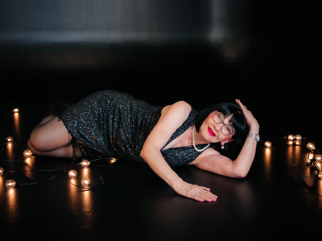 TiniaMartin lies on black floor, wearing sparkly black dress, garter, and stockings, with string lights, red lipstick, glasses, and pearl necklace, arm raised, legs slightly bent.