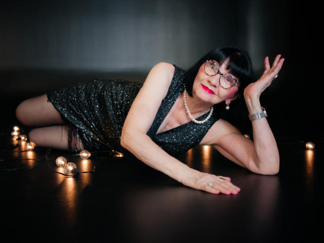 TiniaMartin lying on black floor, wearing sparkly black dress, pearl necklace, glasses, red lipstick, black bob, and bracelet, with candles around, hand on forehead, looking at camera.