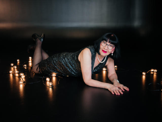TiniaMartin is lying on her stomach, wearing a sparkly black dress, glasses, and a pearl necklace, with Christmas lights around her. She has red lipstick and black heels.