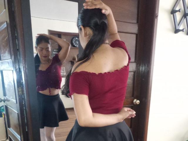 ArianithaXox with long black hair in a ponytail, facing mirror, wearing red off-shoulder top, black skirt, and white socks, showing her back and small ass.