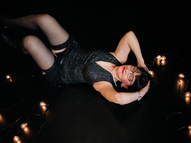 TiniaMartin lies on black floor, arms above head, wearing sparkly black dress, black shorts, sheer stockings, and boots. Glowing lights surround her. She has red lipstick and glasses.