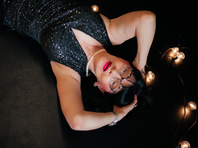 TiniaMartin laying on black floor, arms above head, wearing black sequin dress, pearl necklace, red lipstick, glasses, and black hair, with string of lights to the right.