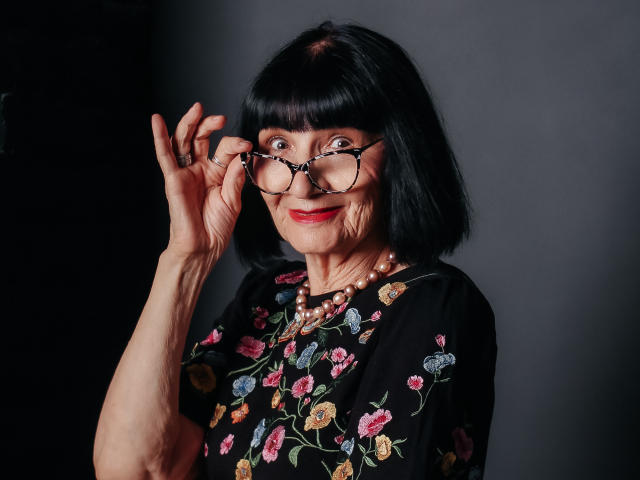 TiniaMartin with black bob, wearing floral black top, holding glasses, smiling, red lipstick, pearl necklace, dark background, right arm raised, looking through glasses, mid-shot, elegant.